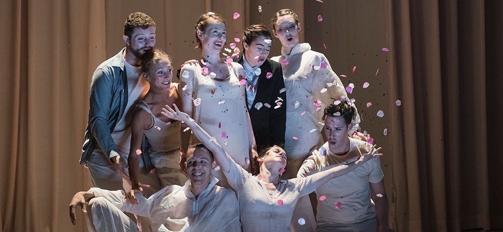Performers pose as a group on stage while one of them throws pink flower petal confetti