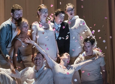 Performers pose as a group on stage while one of them throws pink flower petal confetti