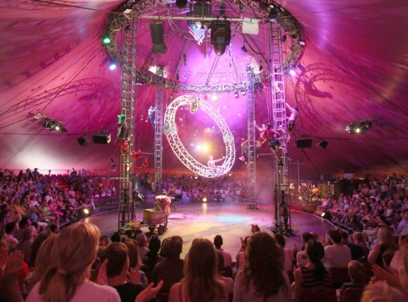 <p>Wide shot of a circus big top tent interior bathed in pink light, showing performers climbing rigging towers and a large illuminated ring suspended overhead, with a packed audience seated in the round.</p>