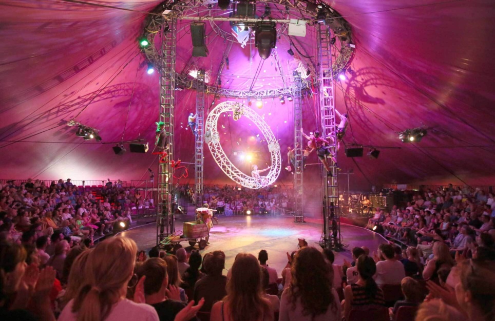 <p>Wide shot of a circus big top tent interior bathed in pink light, showing performers climbing rigging towers and a large illuminated ring suspended overhead, with a packed audience seated in the round.</p>