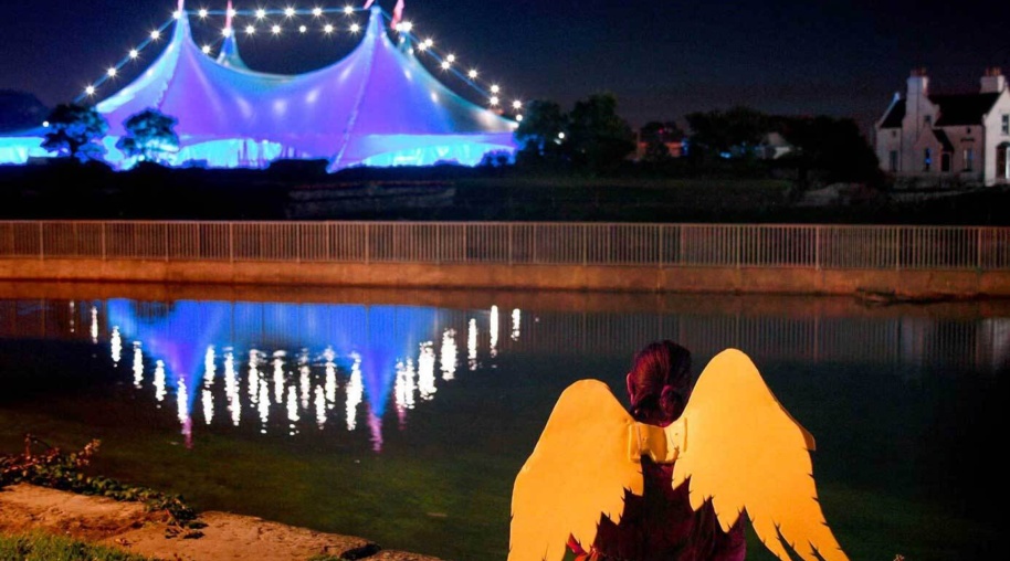 <p>Galway international arts festival, girl with wings witting beside a river looking at the big top tents</p>