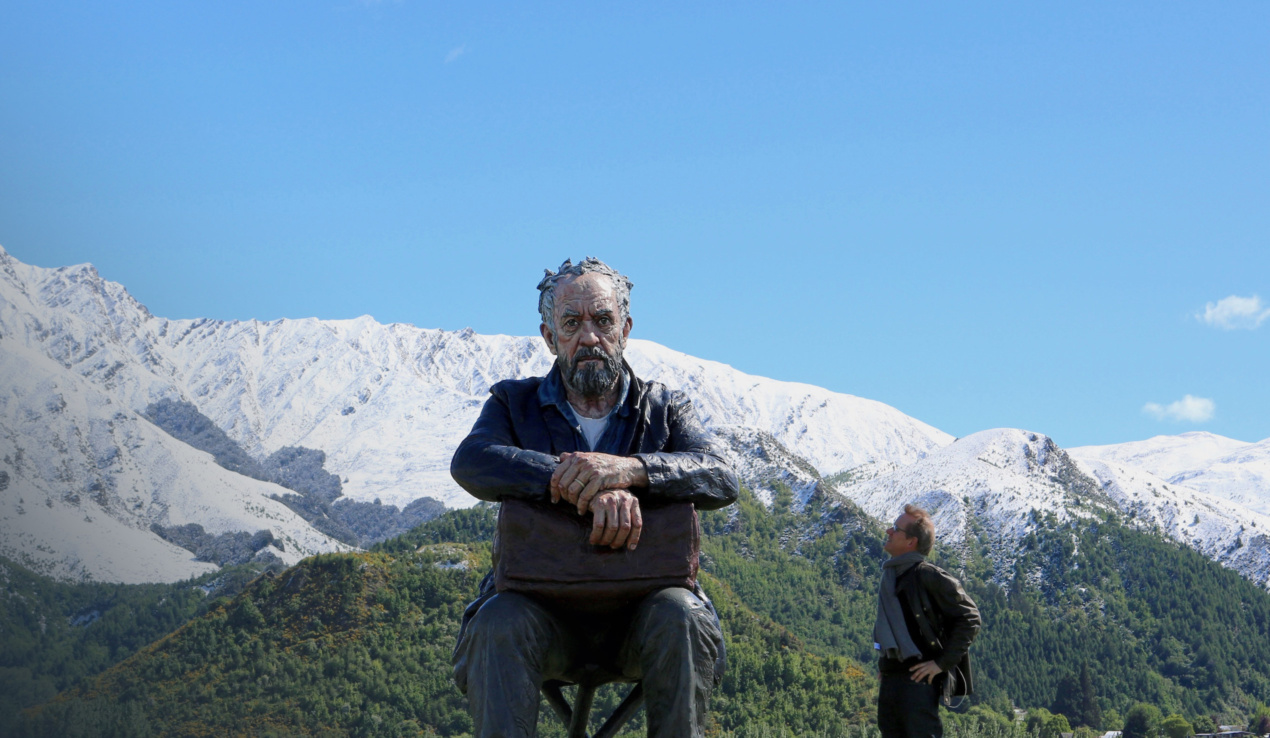 <p>Large sculpture of an old man sitting. A real man looks up at him. Behind them are green fields, snowy mountains, and a clear blue sky.</p>