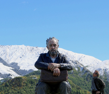 <p>Large sculpture of an old man sitting. A real man looks up at him. Behind them are green fields, snowy mountains, and a clear blue sky.</p>