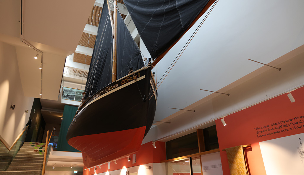 A traditional Galway hooker boat with red hull and black sails suspended in the air in a museum building
